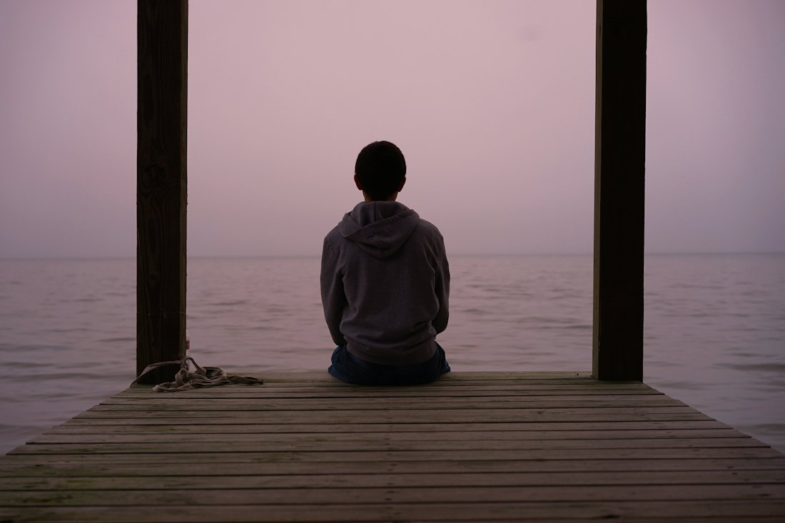 A person sitting alone on a dock at dusk, looking over calm water